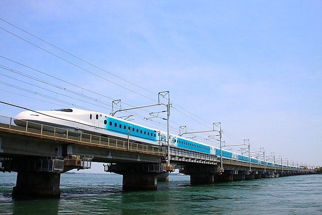 The blue-and-white Sparkling Dreams Shinkansen crossing a bridge over water beneath a blue sky