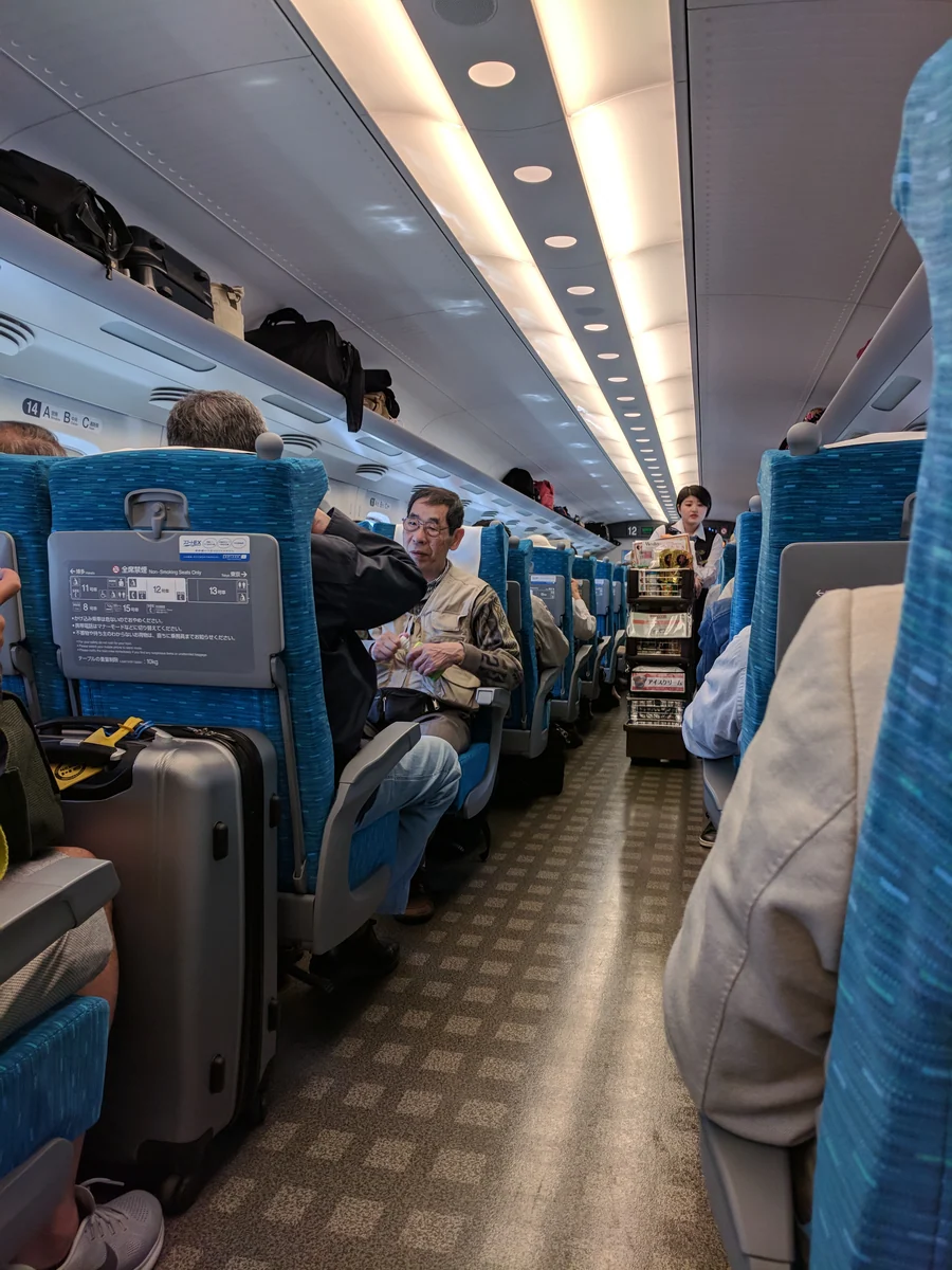 A uniformed attendant with a refreshments trolley among blue seats in the Shinkansen aisle near Odawara, photographed in April 2018.