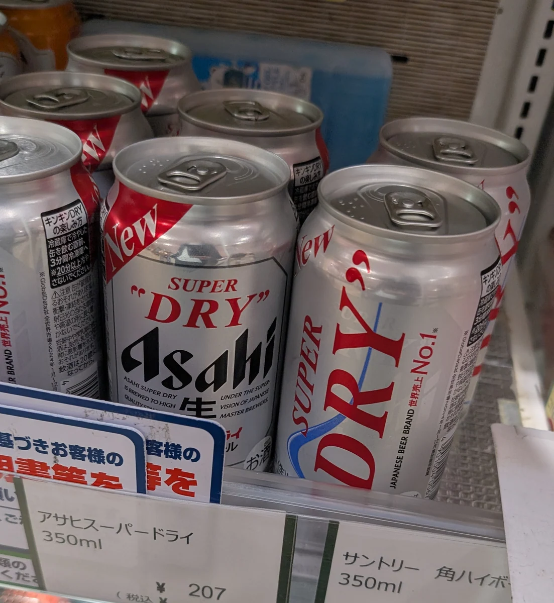 Silver Asahi Super Dry cans on a refrigerated shop shelf, with SUPER DRY, Asahi and 生 visible.