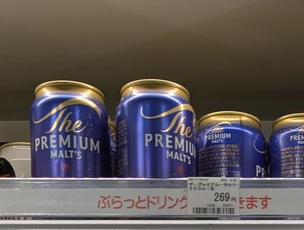 Blue cans of Suntory The Premium Malt’s with gold lettering on the top shelf of a drinks display.