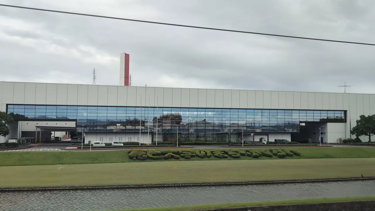 A long white factory beyond a lawn, with the Shinkansen reflected along the bottom of its glass
